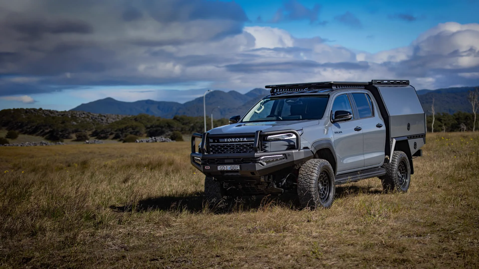 Grey Toyota HiLux with black bull bar, roof rack and custom Camesco aluminium canopy parked in mountain grassland